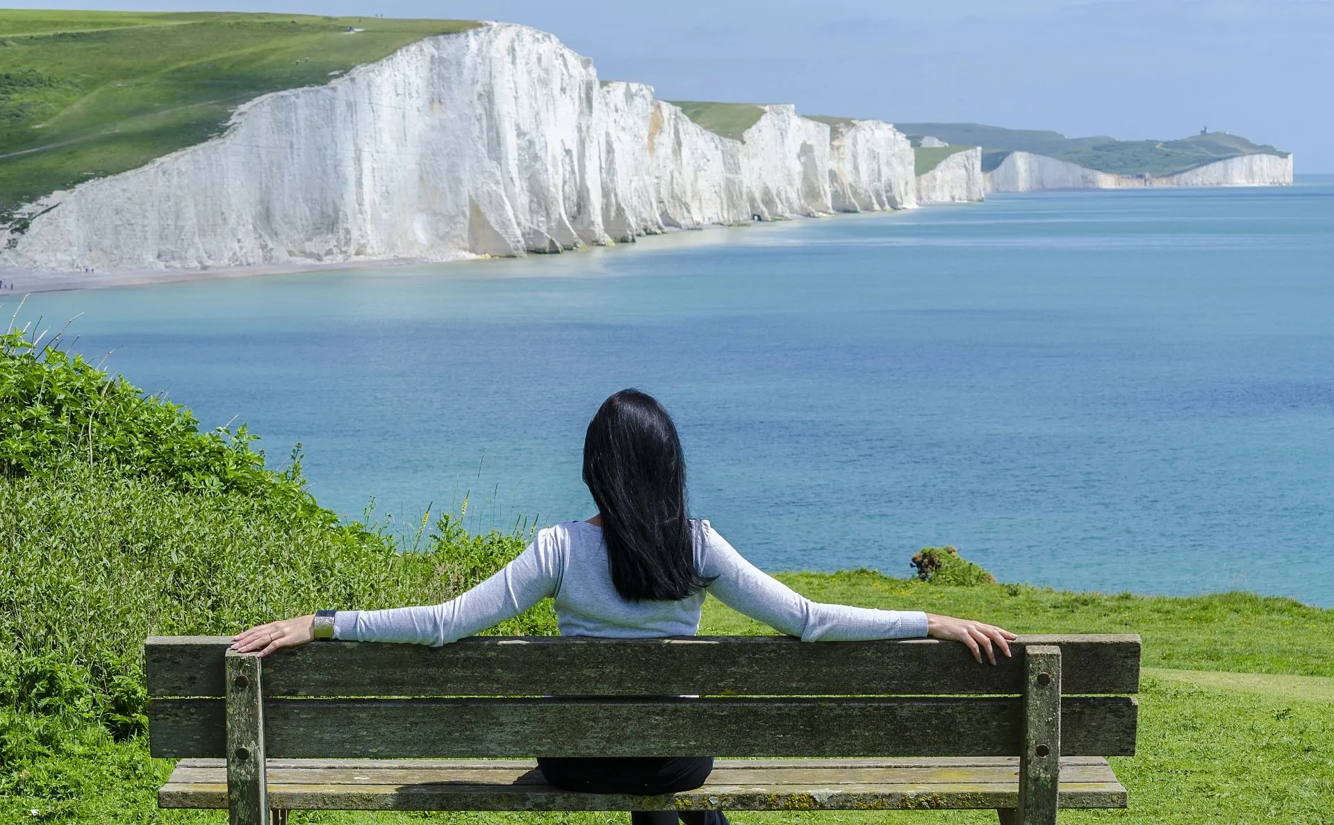 Woman sitting on bench in front of beach with a cliff side in the background