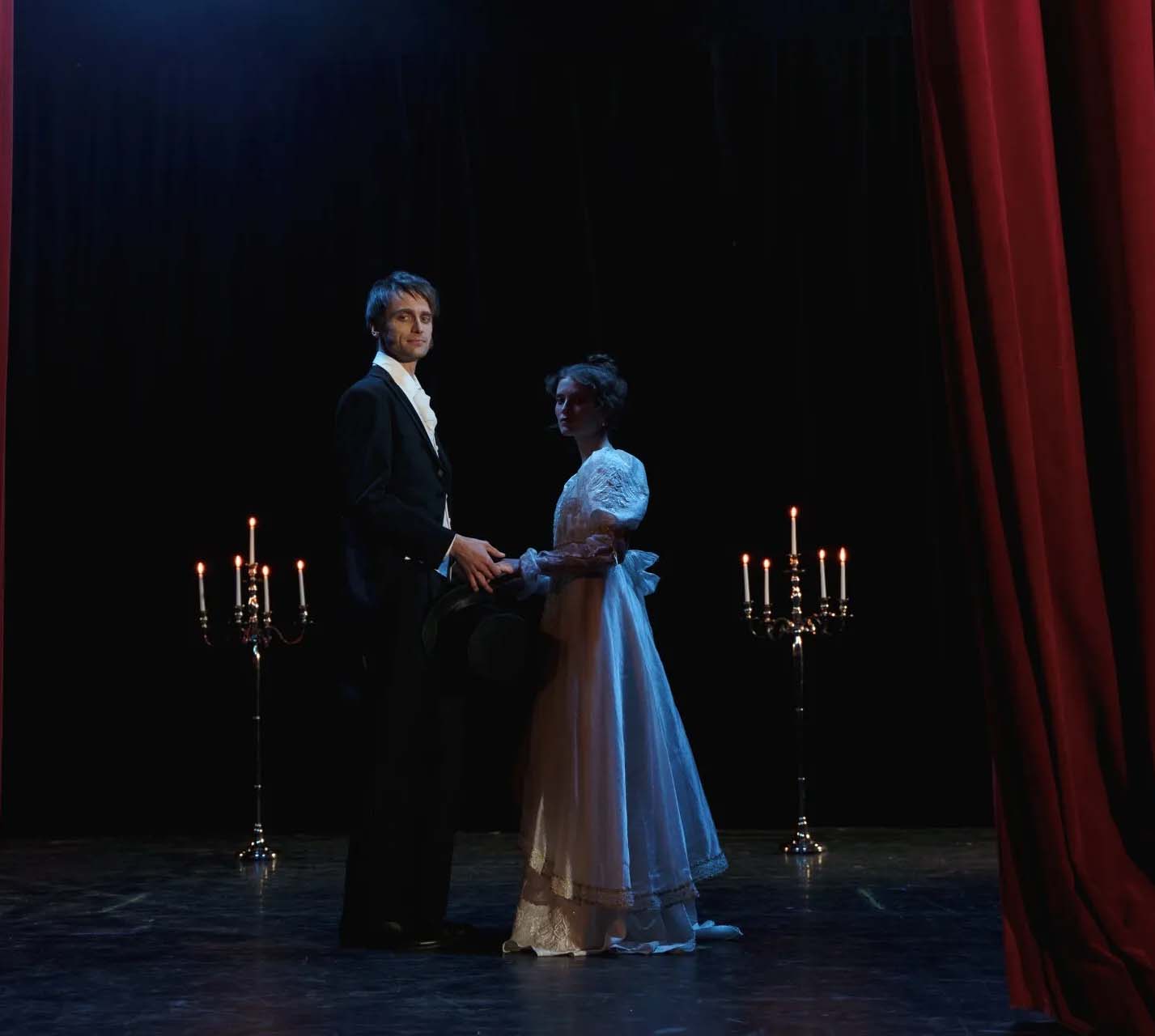 Actor and actress in mid-century costumes on a dark stage with candles lit in the background