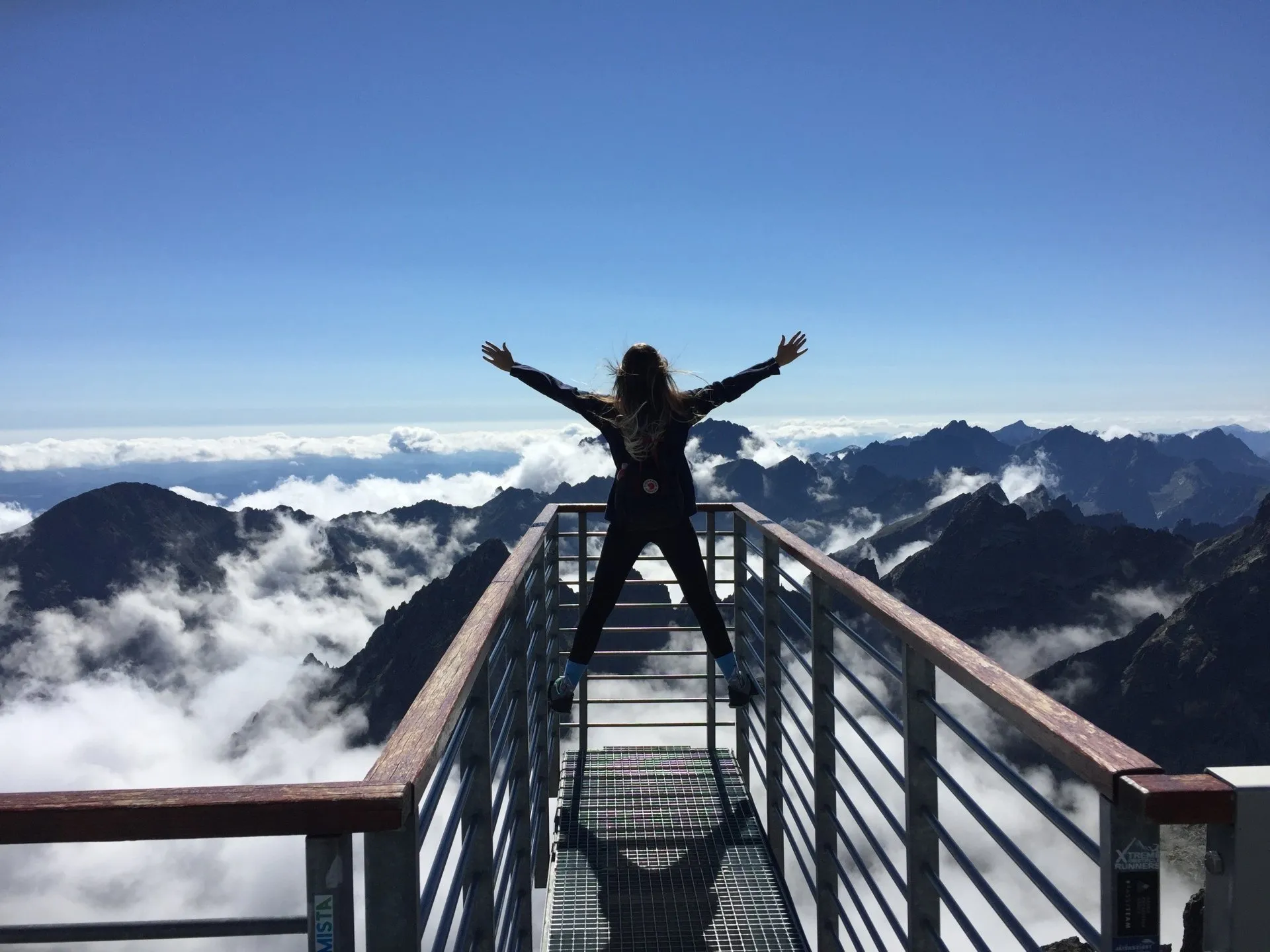 Woman standing on bars of a walkway overlooking mountains and clouds below her with her arms spread wide